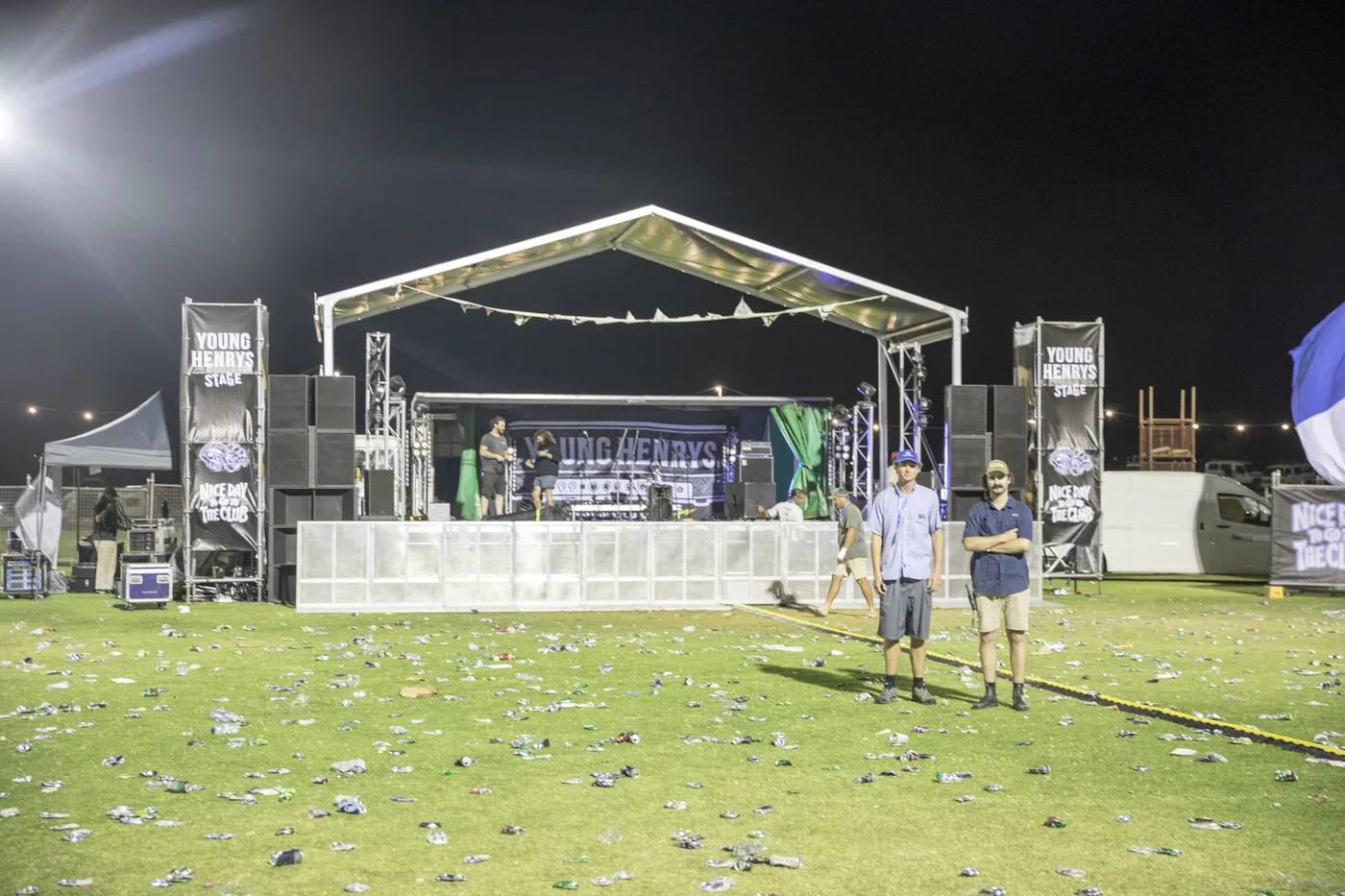 Tom and Chris standing on the empty oval at night, rubbish everywhere, the stage lit up behind them